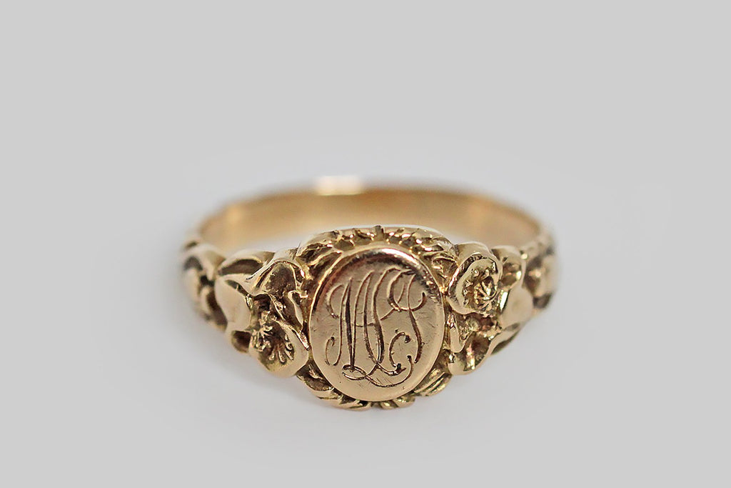 An antique, Art Nouveau era signet ring, made in 14k yellow gold, with ornate, carved details that are especially well-preserved. The ring’s shoulders are adorned with a pair of realistic, fleshy poppies, whose toothed leaves travel down the ring shank to the base, where their stems twine together. Its round face is engraved with the monogram MLJ, in curling period script. 