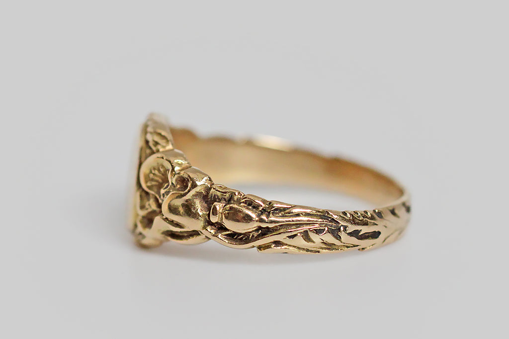 An antique, Art Nouveau era signet ring, made in 14k yellow gold, with ornate, carved details that are especially well-preserved. The ring’s shoulders are adorned with a pair of realistic, fleshy poppies, whose toothed leaves travel down the ring shank to the base, where their stems twine together. Its round face is engraved with the monogram MLJ, in curling period script. 