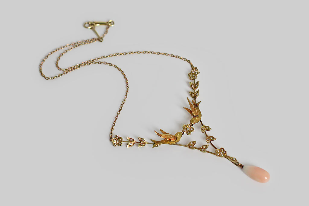A Victorian-era necklace, with a sweet, serene presence. At its center, a pair of three-dimensional swallows come together to hold a five-petaled, pearl-set flower. An elegant, v-shaped framework of flowers and leaves is built beneath these bird friends, and a pair of jointed flowers attach them, from above, to the necklace's fine cable chain. All of the floral elements are set with natural seed pearls.