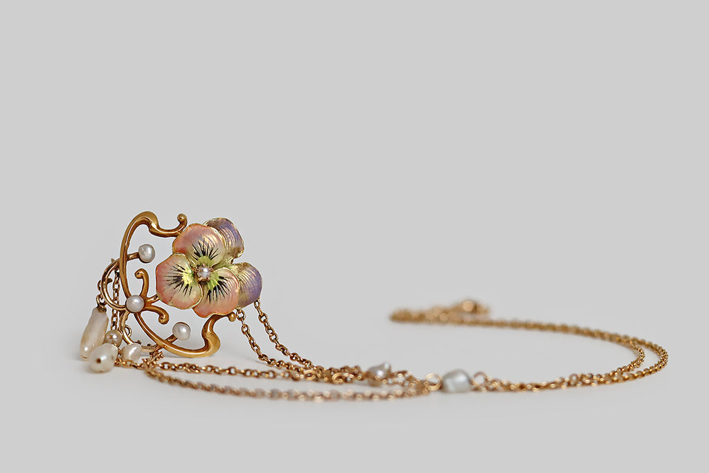 Poor Mouchette | Curated Antique Jewelry, Vintage Jewelry & Engagement Rings | Portland, Oregon | A beautiful, Art Nouveau era festoon necklace, modeled in rosy 14k yellow gold, whose focal point is a cheerful pansy blossom, decorated with iridescent, multi-color enamel. This sweet, pastel pansy rests inside a shapely Art Nouveau frame. White seed pearls decorate the frame, while a trio of natural river pearls dangle below it; further small pearls decorate the necklace’s chain