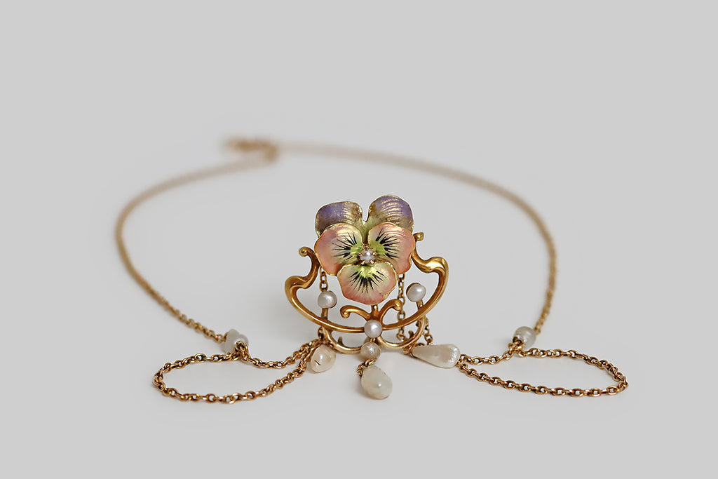 Poor Mouchette | Curated Antique Jewelry, Vintage Jewelry & Engagement Rings | Portland, Oregon | A beautiful, Art Nouveau era festoon necklace, modeled in rosy 14k yellow gold, whose focal point is a cheerful pansy blossom, decorated with iridescent, multi-color enamel. This sweet, pastel pansy rests inside a shapely Art Nouveau frame. White seed pearls decorate the frame, while a trio of natural river pearls dangle below it; further small pearls decorate the necklace’s chain