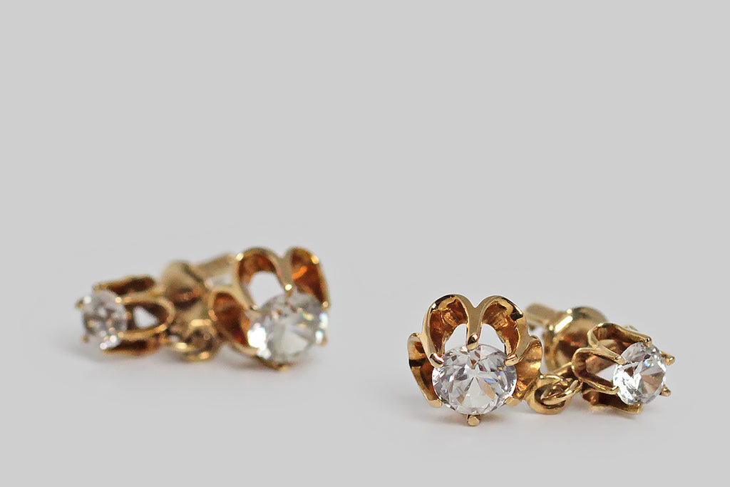 A pair of late-Victorian drop earrings, modeled in 14k yellow gold, whose buttercup mounts hold a plump suite of sparkling paste stones. These vibrant old imposters look very much like real diamonds, thanks to their hand-cut nature. The buttercup settings are designed with dramatic, rounded, cutaway sections, and six claw-like prong that hold the paste gems aloft. These earrings are short drops