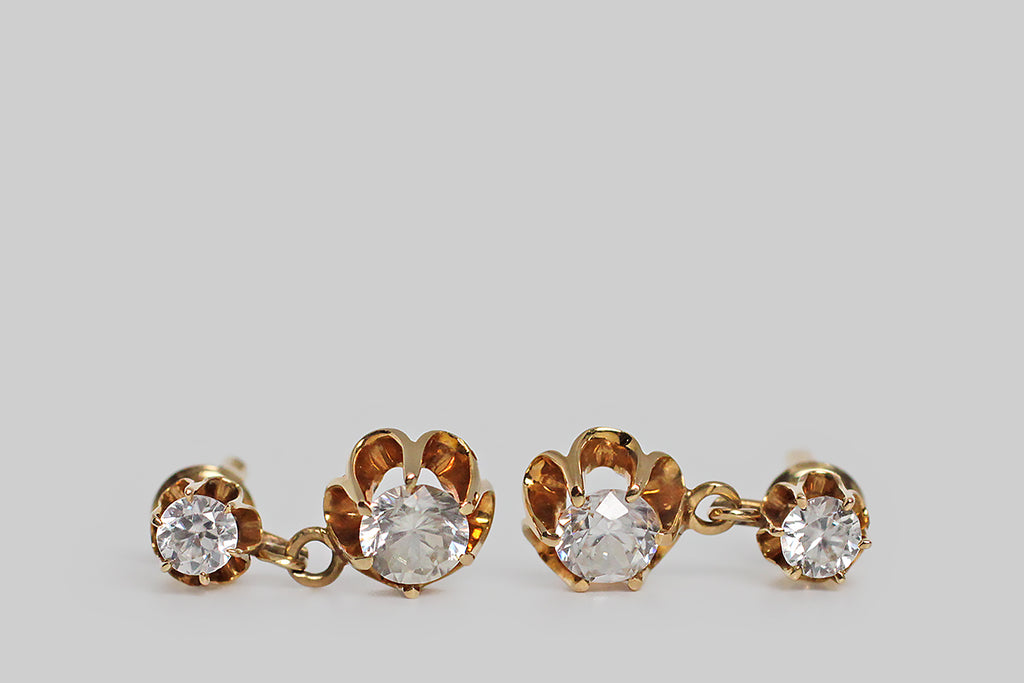 A pair of late-Victorian drop earrings, modeled in 14k yellow gold, whose buttercup mounts hold a plump suite of sparkling paste stones. These vibrant old imposters look very much like real diamonds, thanks to their hand-cut nature. The buttercup settings are designed with dramatic, rounded, cutaway sections, and six claw-like prong that hold the paste gems aloft. These earrings are short drops