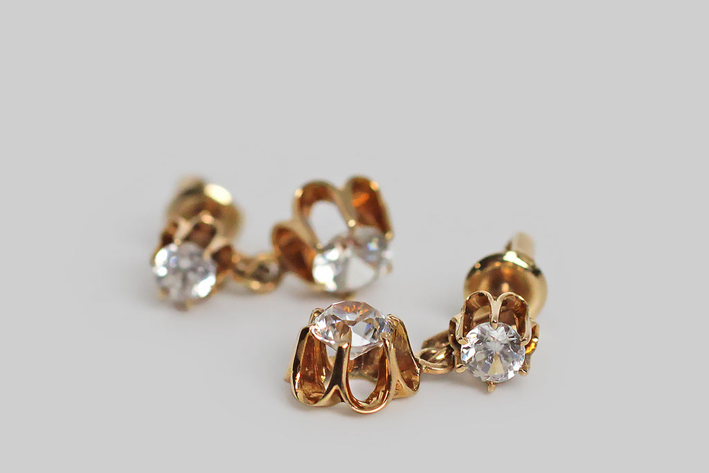A pair of late-Victorian drop earrings, modeled in 14k yellow gold, whose buttercup mounts hold a plump suite of sparkling paste stones. These vibrant old imposters look very much like real diamonds, thanks to their hand-cut nature. The buttercup settings are designed with dramatic, rounded, cutaway sections, and six claw-like prong that hold the paste gems aloft. These earrings are short drops
