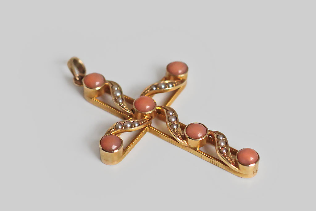 A late-Victorian, cross-shaped pendant, made in 14k yellow gold, embellished with natural coral cabochons and a bevy natural, silvery, seed pearls. This cross is beautifully made, with an airy, open-work character. Its six, pinkish-orange corals are set in smooth bezels— ribbon like forms weave between the coral stations, and each of these is bead-set with three seed pearls. The graceful movement of these ribboning forms calls to mind the image of a cloth draped cross, 