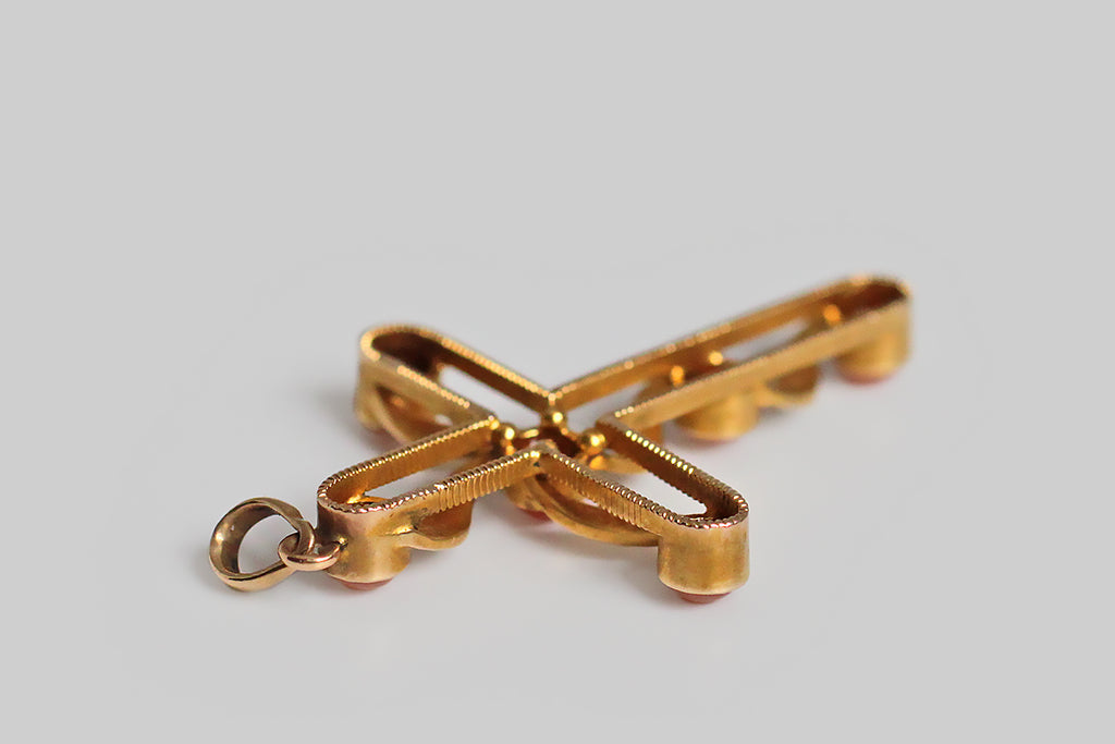 A late-Victorian, cross-shaped pendant, made in 14k yellow gold, embellished with natural coral cabochons and a bevy natural, silvery, seed pearls. This cross is beautifully made, with an airy, open-work character. Its six, pinkish-orange corals are set in smooth bezels— ribbon like forms weave between the coral stations, and each of these is bead-set with three seed pearls. The graceful movement of these ribboning forms calls to mind the image of a cloth draped cross, 