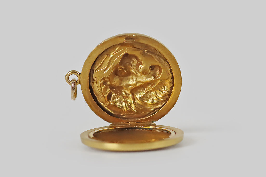 Poor Mouchette | Curated Antique Jewelry, Vintage Jewelry & Engagement Rings | Portland, Oregon | A wonderful Art Nouveau era, repoussé locket, modeled in 14k yellow gold, whose fierce subject is a lion sitting in profile amid a clutch of tall grasses. Our lion is realistically rendered, with a snarling, focused expression; his long, wavy mane mingles with the graceful blades of grass that surround him. He holds a sparkling, prong-set, old mine cut diamond in his mouth.